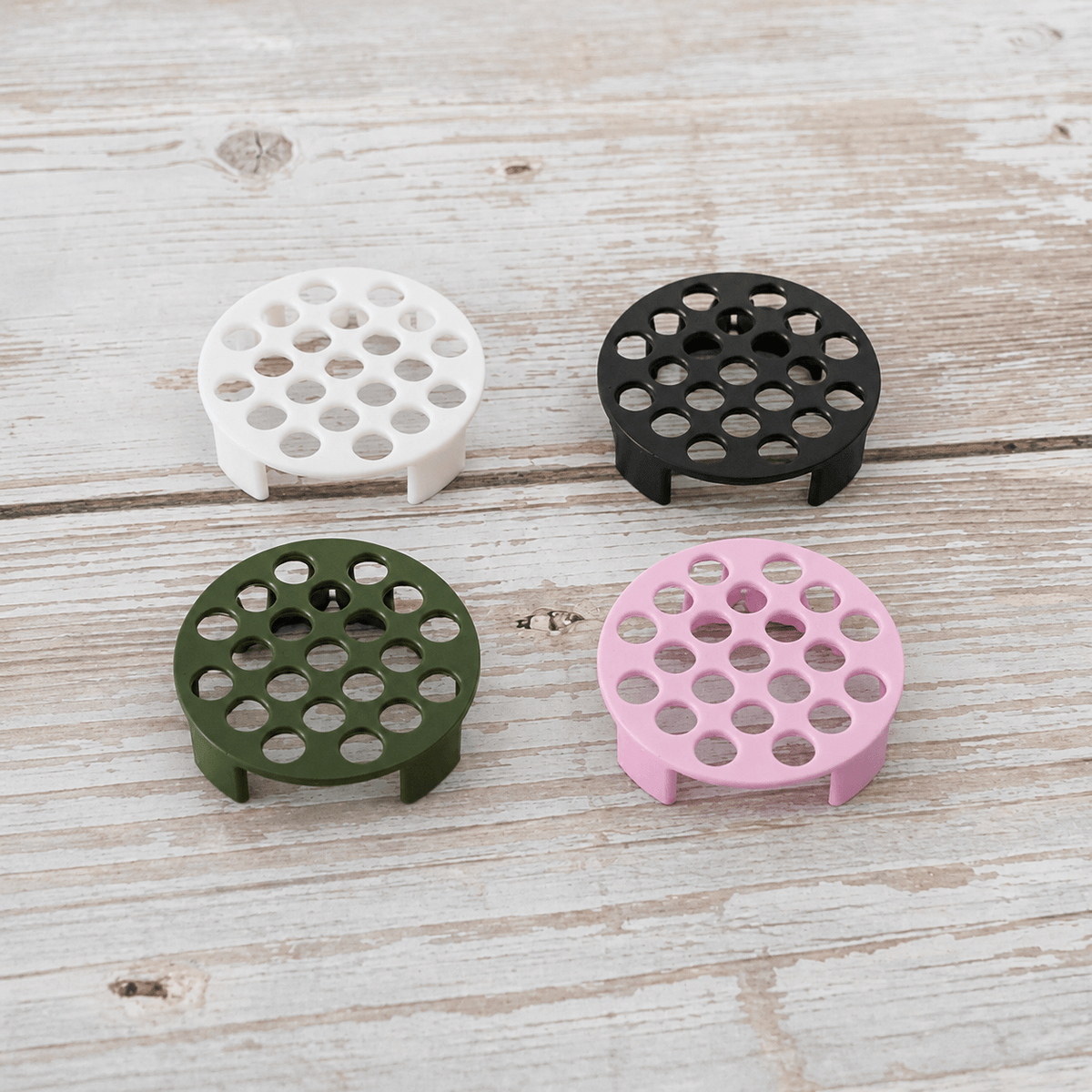 Four round plastic stand risers with circular cutouts, shown in white, black, olive green, and light pink on a whitewashed wooden surface.