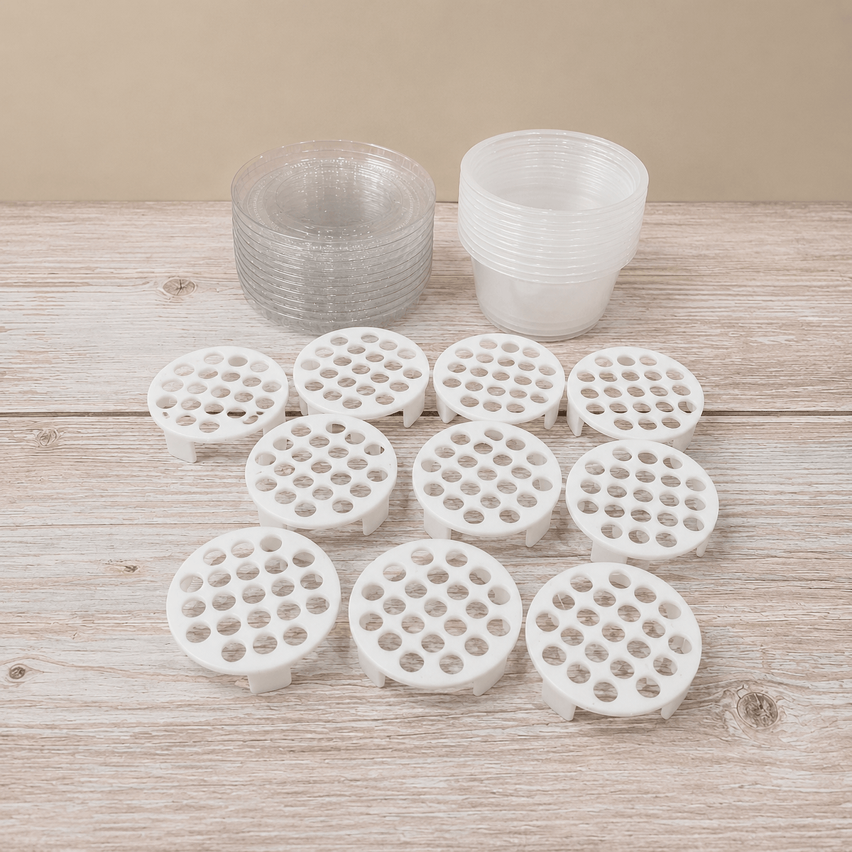 White circular plastic strainer baskets with multiple drainage holes, arranged on a light wood surface, with two stacks of clear plastic cups behind them.