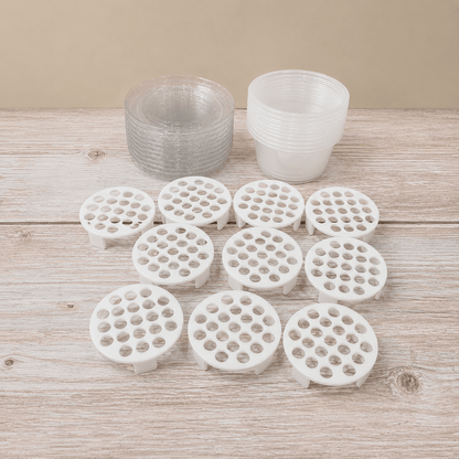 White circular plastic strainer baskets with multiple drainage holes, arranged on a light wood surface, with two stacks of clear plastic cups behind them.