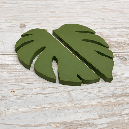 A pair of olive green wooden monstera leaf cutouts displayed on a light, rustic white-washed wooden surface.