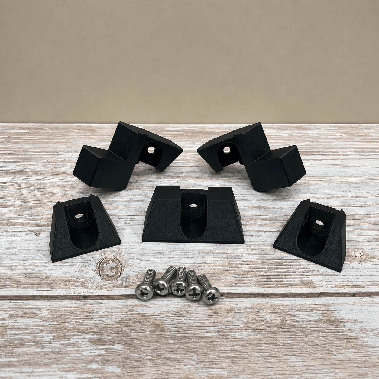 Five black plastic mounting brackets with silver screws, arranged on a rustic white-washed wooden surface against a solid beige background.