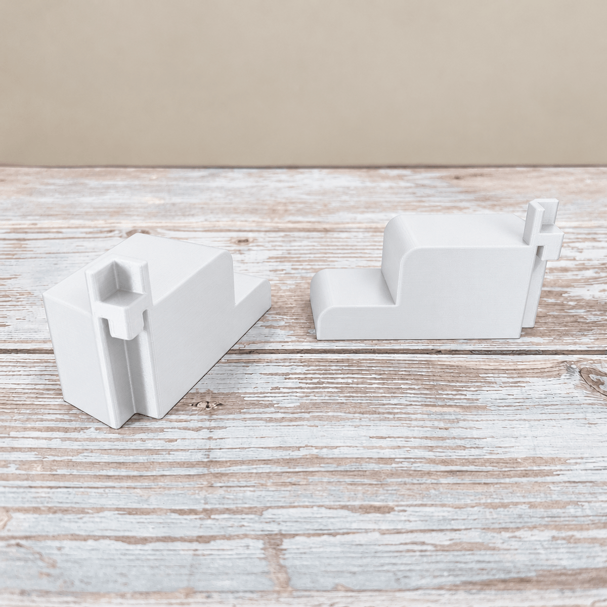 Two white PETG wire shelf brackets for an IKEA Rudsta cabinet, showing the mounting channels and side profile on a whitewashed wood surface.