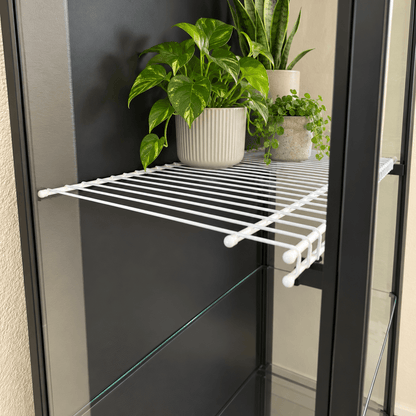 Side-angle view of a white wire shelf with black mounting brackets in an IKEA Rudsta cabinet, with potted plants toward the back.