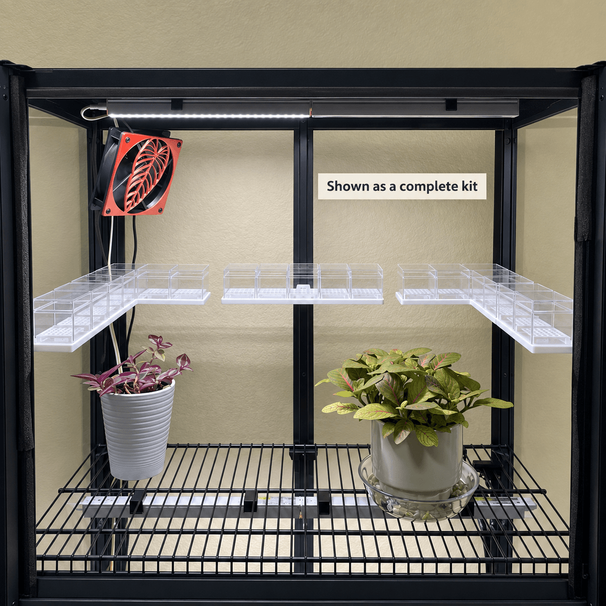 A black metal grow light kit with two shelves, clear plastic plant holders, a hanging fan, and a light bar, shown with two potted plants against a beige wall. A text label reads 'Shown as a complete kit'.