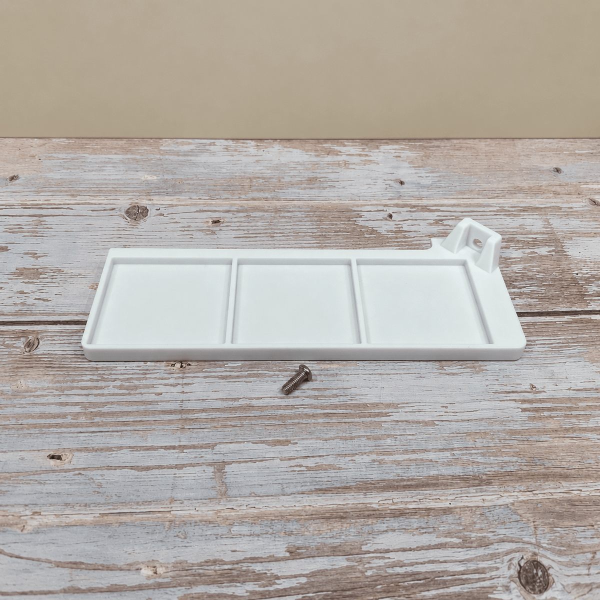 A white three-compartment tray with a small mounting bracket on one end, shown on a rustic whitewashed wooden surface with a small metal screw nearby.