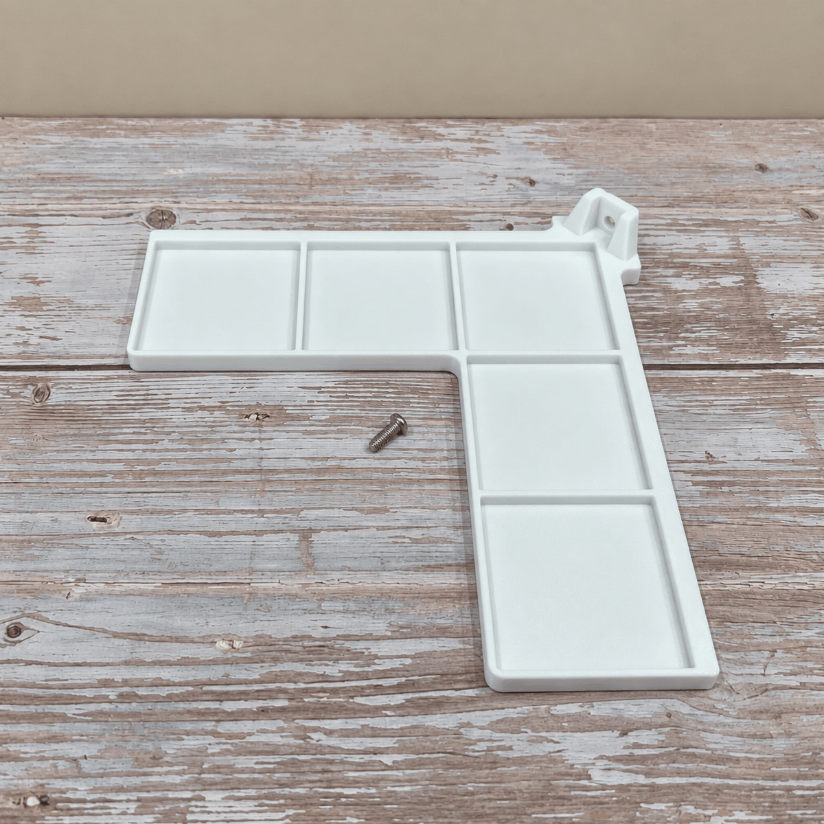 A white plastic L-shaped wall-mounted paint palette with six compartments and a small metal mounting screw, displayed on a rustic wooden surface.