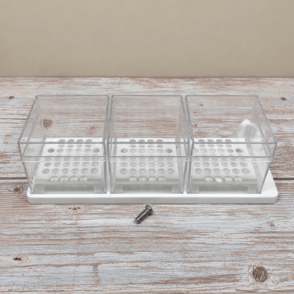 A three-compartment clear plastic organizer tray with a white base, shown on a rustic whitewashed wooden surface against a beige background. A small metal screw is placed in front of the tray.