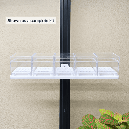 A clear plastic grow tray kit with four compartments mounted on a black vertical support against a beige stucco wall, with a small green plant visible in the lower right corner and a text label reading 'Shown as a complete kit'.
