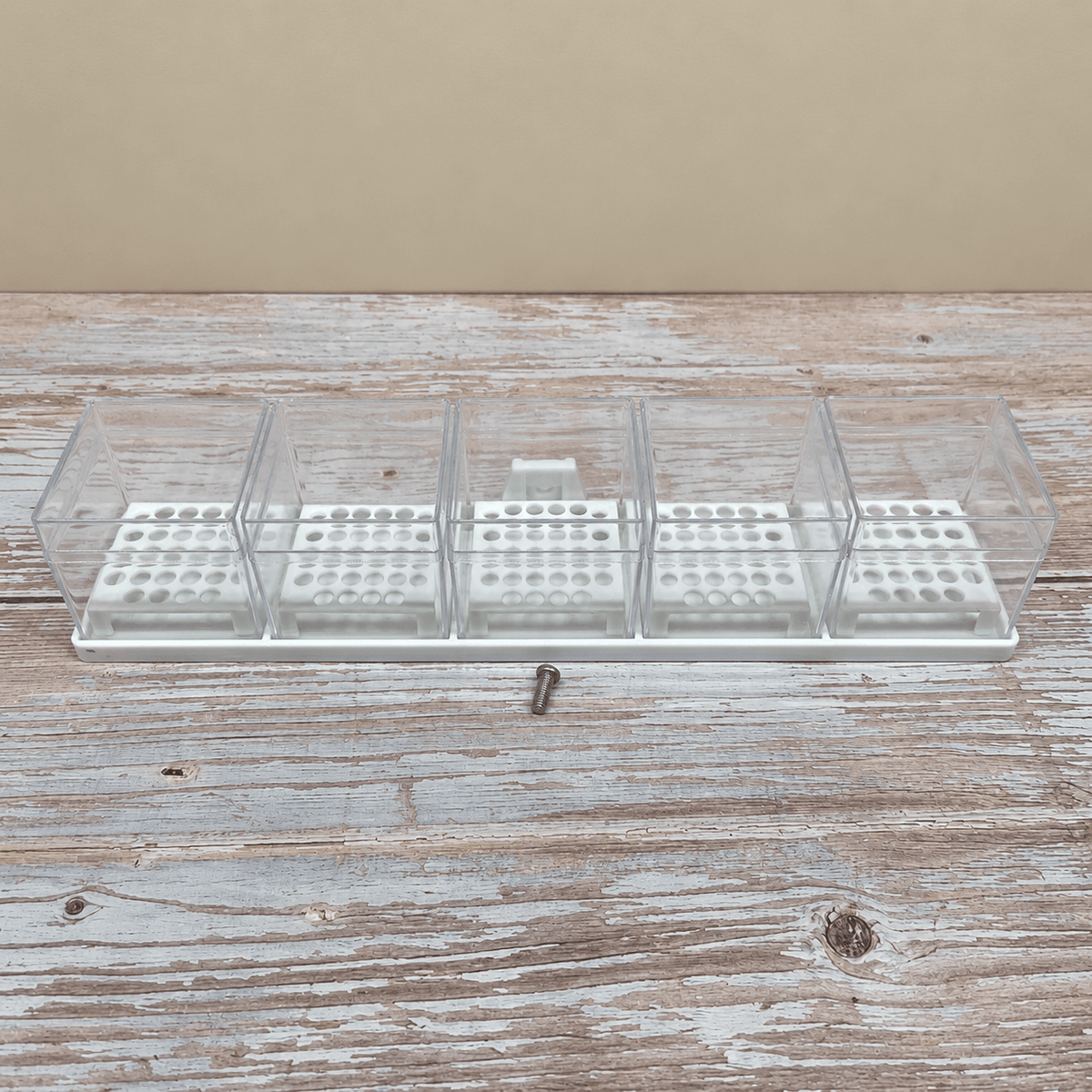A clear plastic multi-compartment organizer tray with a white base, shown on a rustic wooden surface with a small metal screw in front of it.