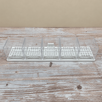 A clear plastic multi-compartment organizer tray with a white base, shown on a rustic wooden surface with a small metal screw in front of it.