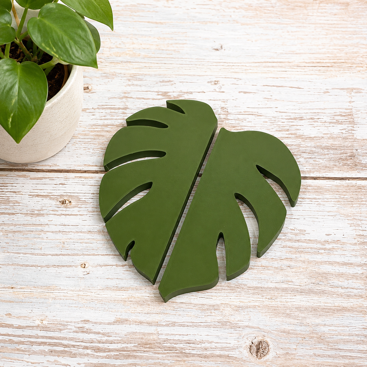 A green wooden monstera leaf-shaped wall decor piece displayed on a light whitewashed wooden surface next to a potted green plant.