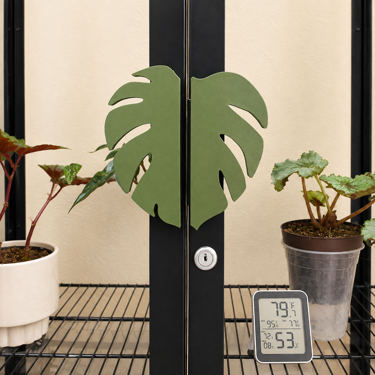 A pair of green monstera leaf-shaped door handles mounted on black metal doors, with potted plants and a digital thermometer on a wire shelf in the foreground.
