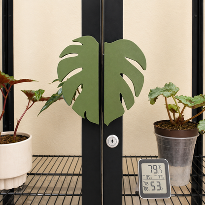 A pair of green monstera leaf-shaped door handles mounted on black metal doors, with potted plants and a digital thermometer on a wire shelf in the foreground.