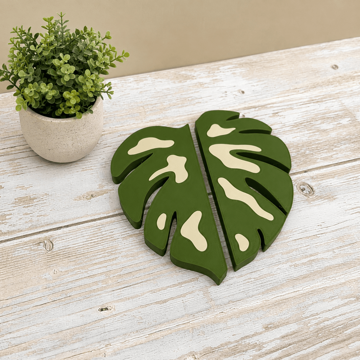A pair of green wooden monstera leaf wall decor pieces with cream-colored accents, displayed on a rustic white-washed wooden table next to a small potted green plant.
