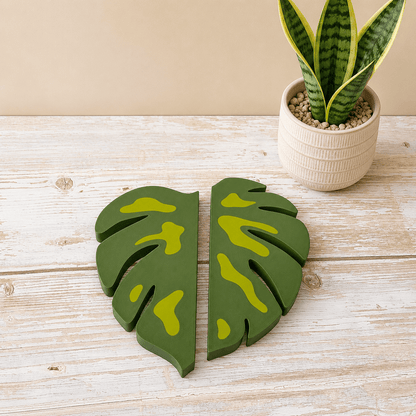 A pair of green monstera leaf-shaped wall decor pieces with yellow-green painted accents, displayed on a light wood tabletop next to a potted snake plant.