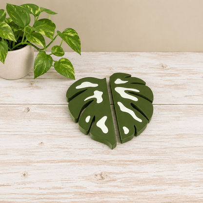 A pair of green monstera leaf-shaped coasters with white leaf veining, displayed on a light wood tabletop next to a potted pothos plant.