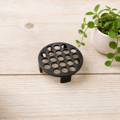 A round black plastic drainage drain cover with multiple circular holes and short legs, shown on a light wood surface next to a potted green plant.