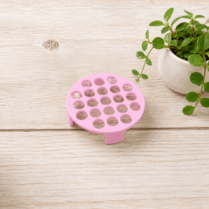 A pink silicone cup lifter with multiple circular holes, shown on a light wood tabletop next to a potted green plant.