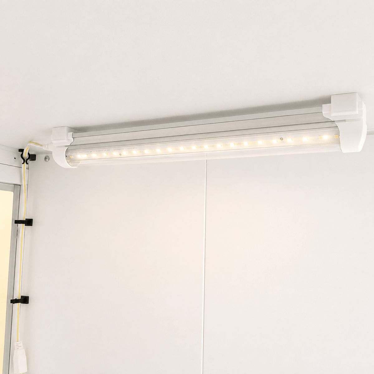 A white LED under-cabinet light bar mounted on a white wall, with its power cord secured on the left using black clips.