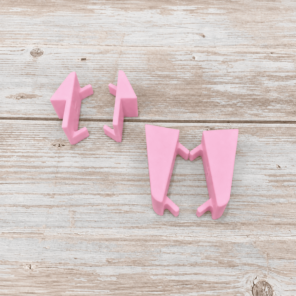 Three pink plastic geometric wall hooks arranged on a light weathered wood surface.