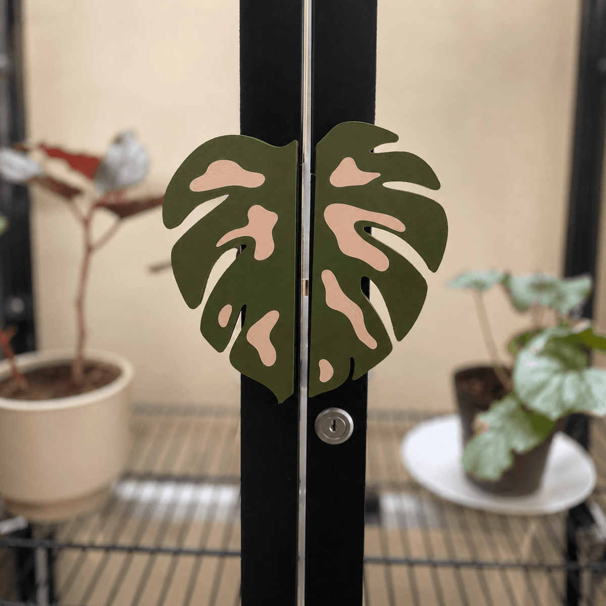 A pair of decorative metal door hinges shaped like split monstera leaves in olive green with pale peach accents, mounted on a black double door in a greenhouse or indoor garden setting with potted plants in the background.