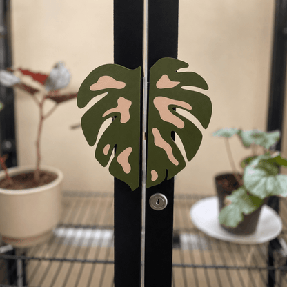 A pair of decorative metal door hinges shaped like split monstera leaves in olive green with pale peach accents, mounted on a black double door in a greenhouse or indoor garden setting with potted plants in the background.
