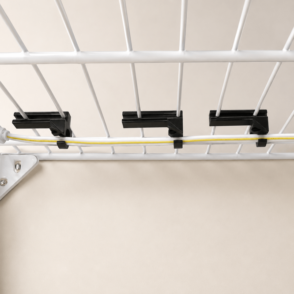 Three black cable clips holding a yellow cable beneath a white wire rack, shown against a plain beige background.