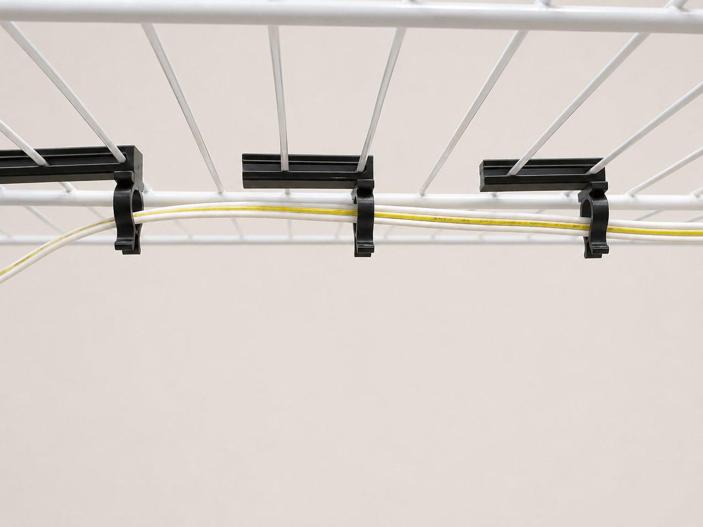 Three black cable management clips holding a yellow cable beneath a white wire rack against a light beige background.