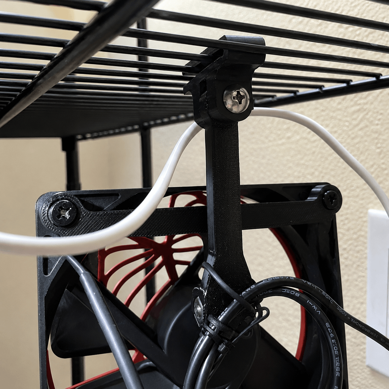 A close-up of a black 3D-printed cable management bracket holding a computer cooling fan, with white and black cables routed underneath a black wire rack.
